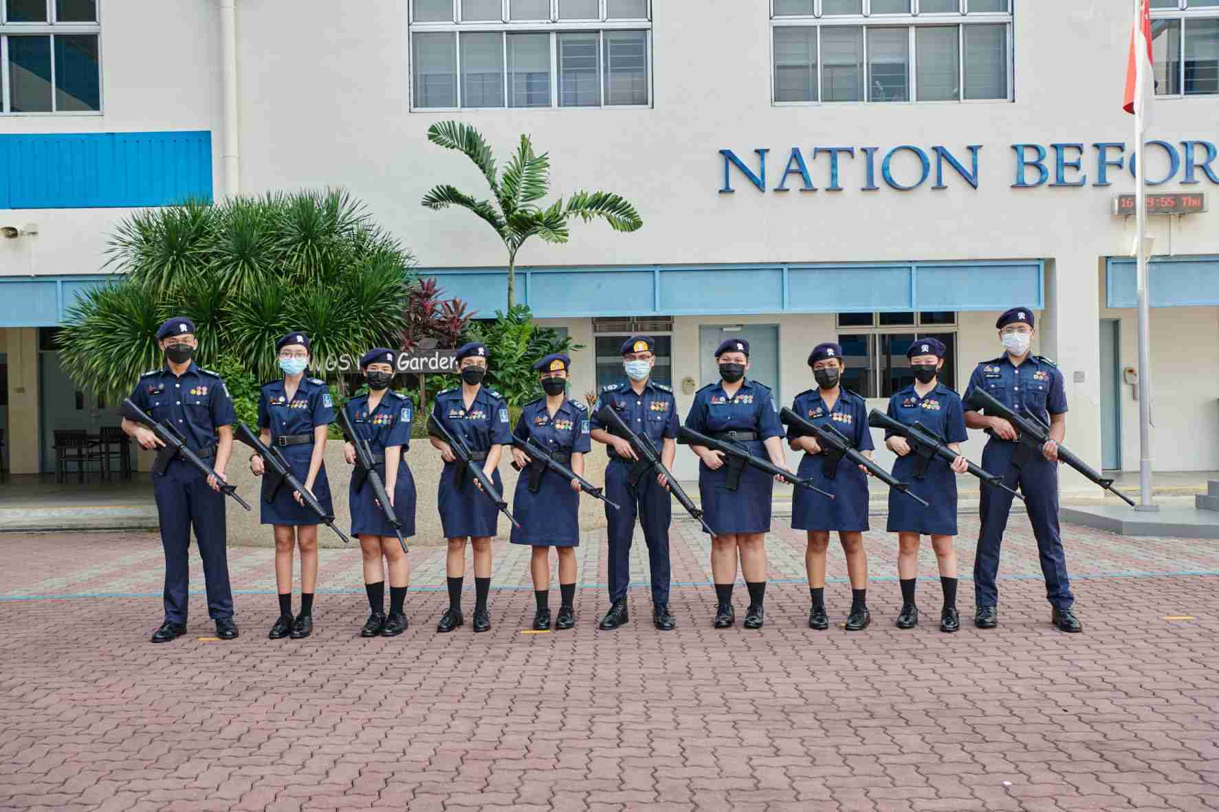 Ten uniformed cadets in navy blue holding rifles stand in a row outside a building reading "Nation Before."